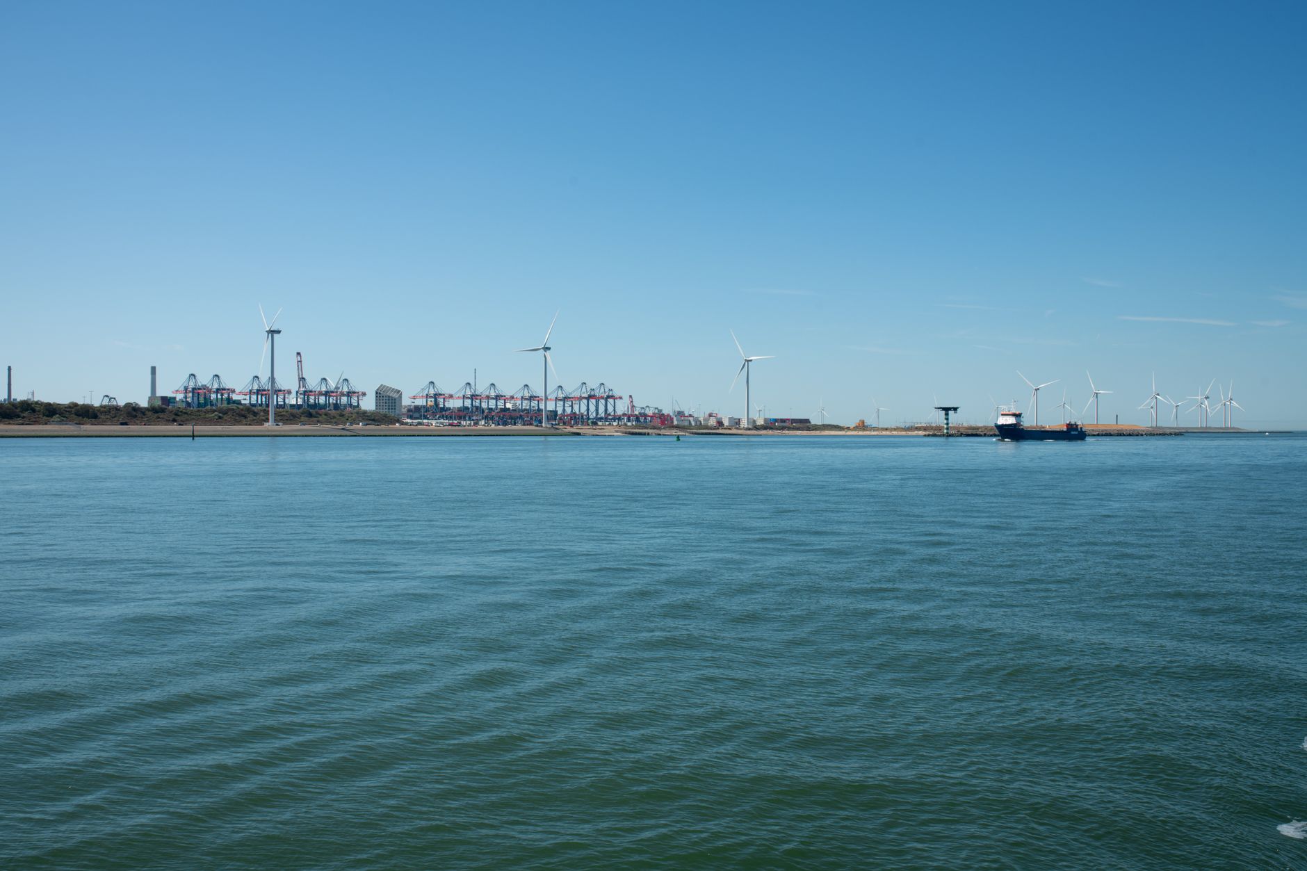 Vue large d’un port maritime avec mer au premier plan, grues à conteneurs, éoliennes et un navire de charge au loin s…
