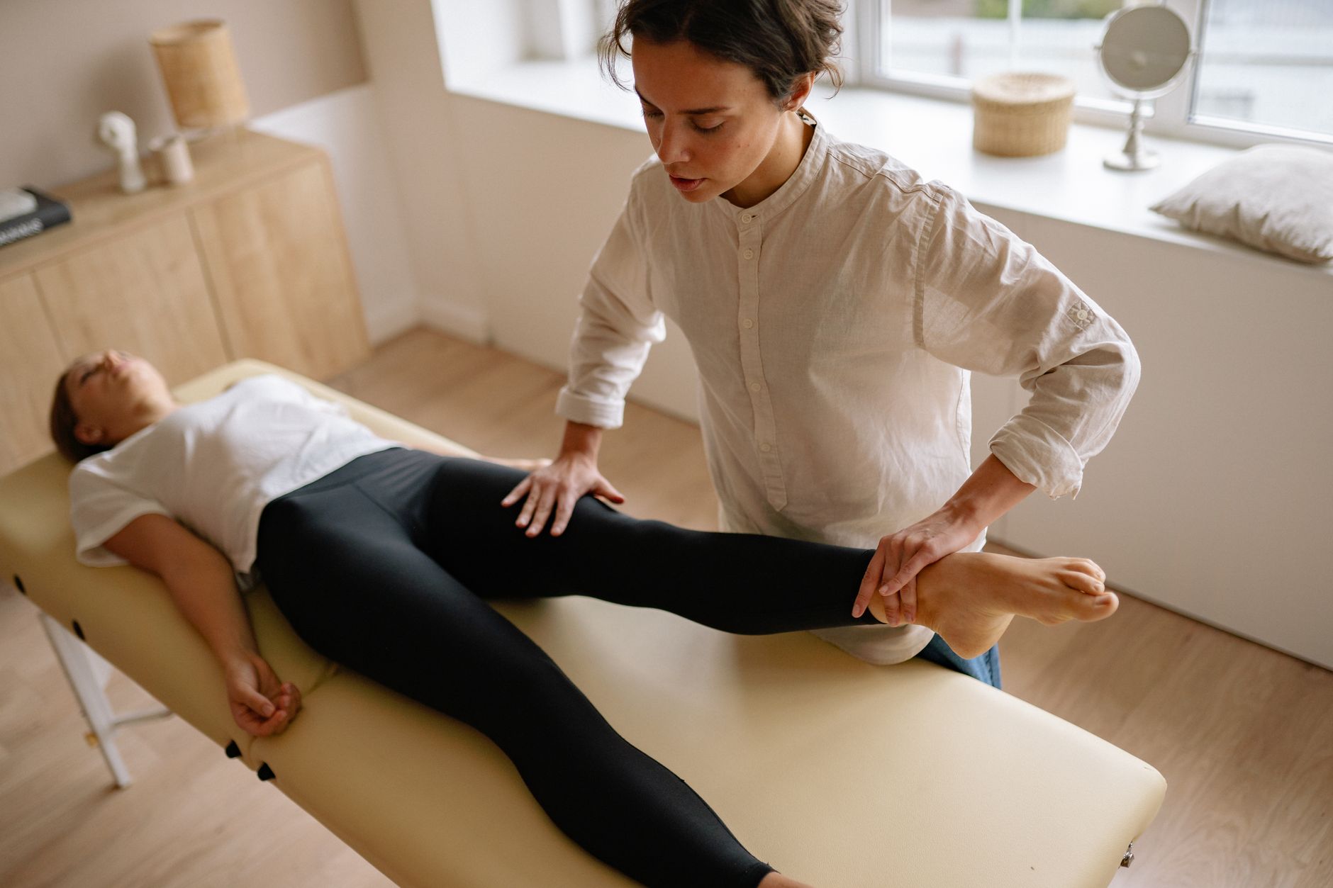 Une praticienne mobilise la jambe d’une personne allongée sur une table de soin, dans un cabinet lumineux.