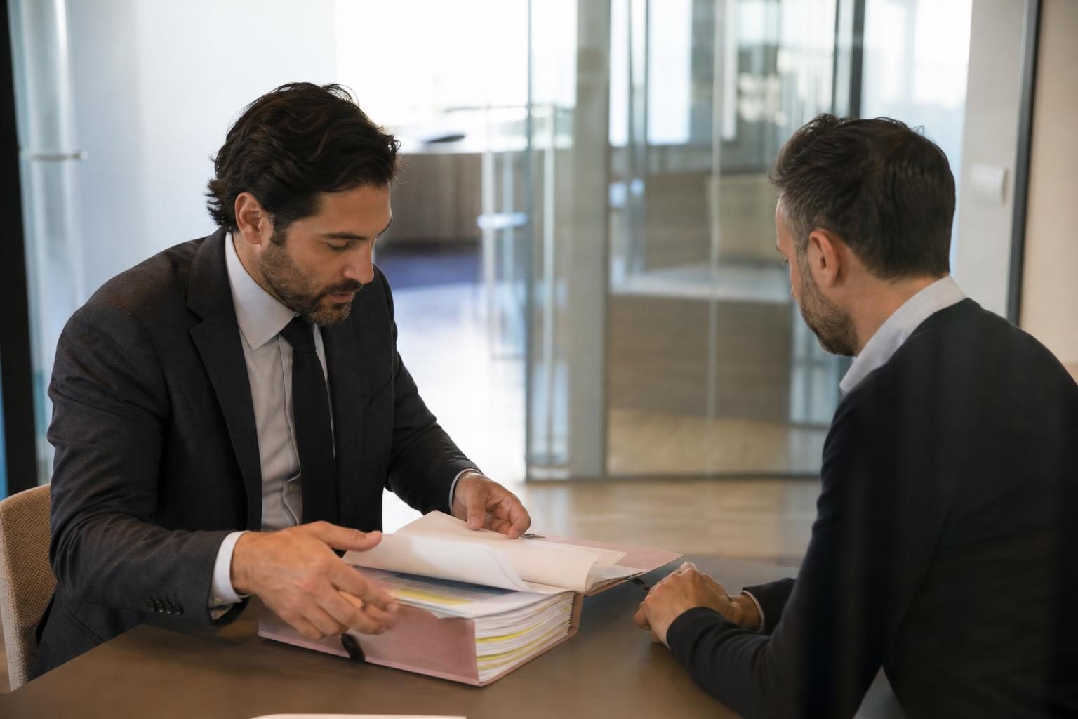 Deux hommes en costume sont assis face à face à une table de bureau, l’un feuillette un gros dossier papier