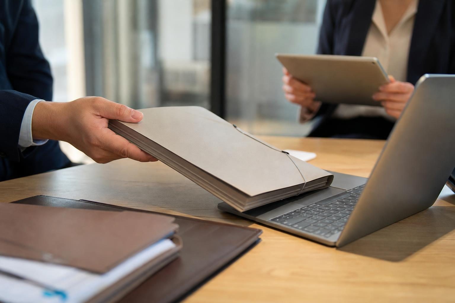 Une personne tend un épais dossier papier au-dessus d’un ordinateur portable, tandis qu’une autre personne tient une tablette en arrière-plan.