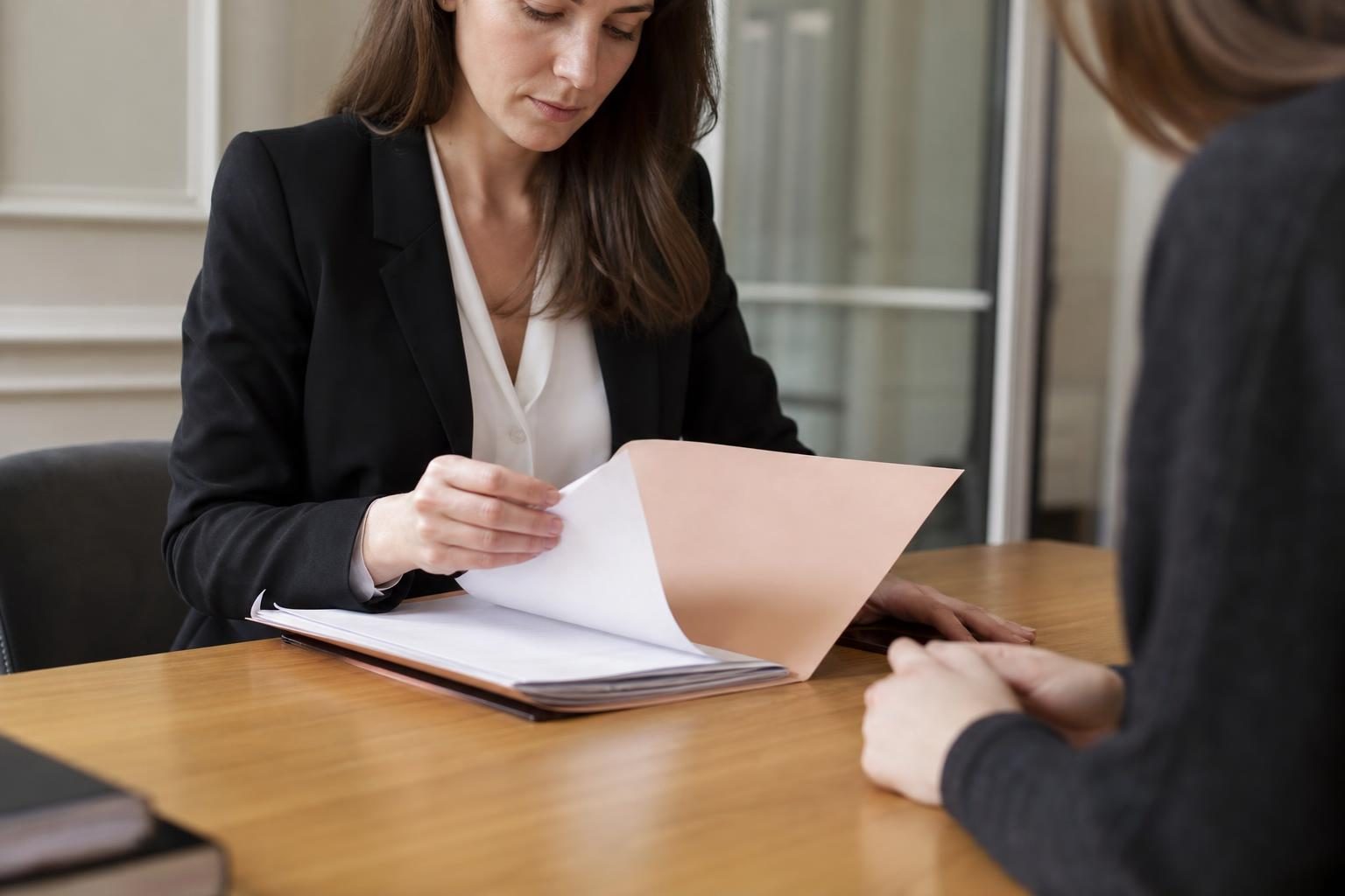 Une avocate en tenue sombre consulte un dossier dans un bureau, face à une cliente partiellement visible de dos.