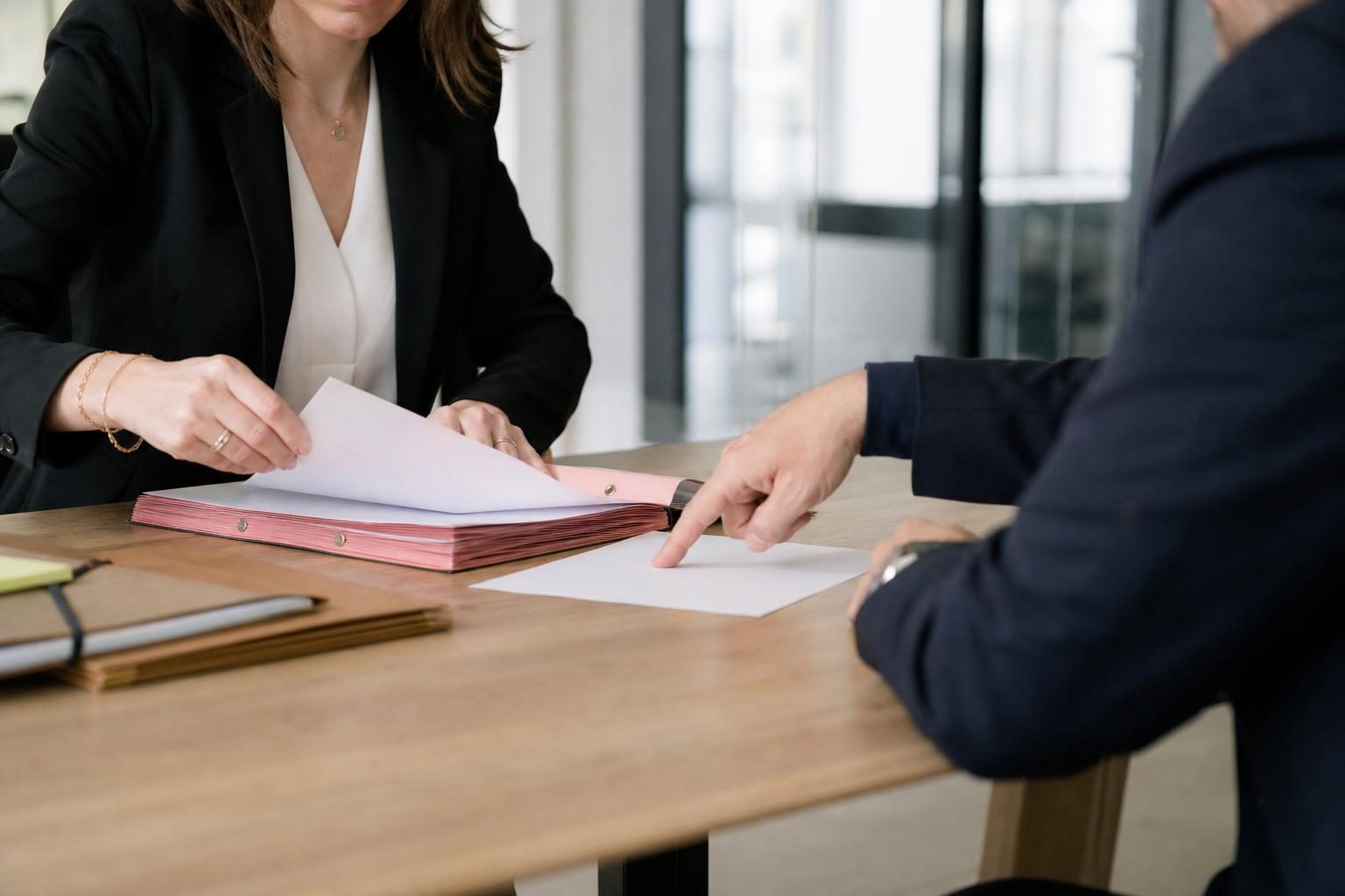 Une avocate en veste sombre consulte un dossier sur une table avec un interlocuteur dont on ne voit que le bras et le…