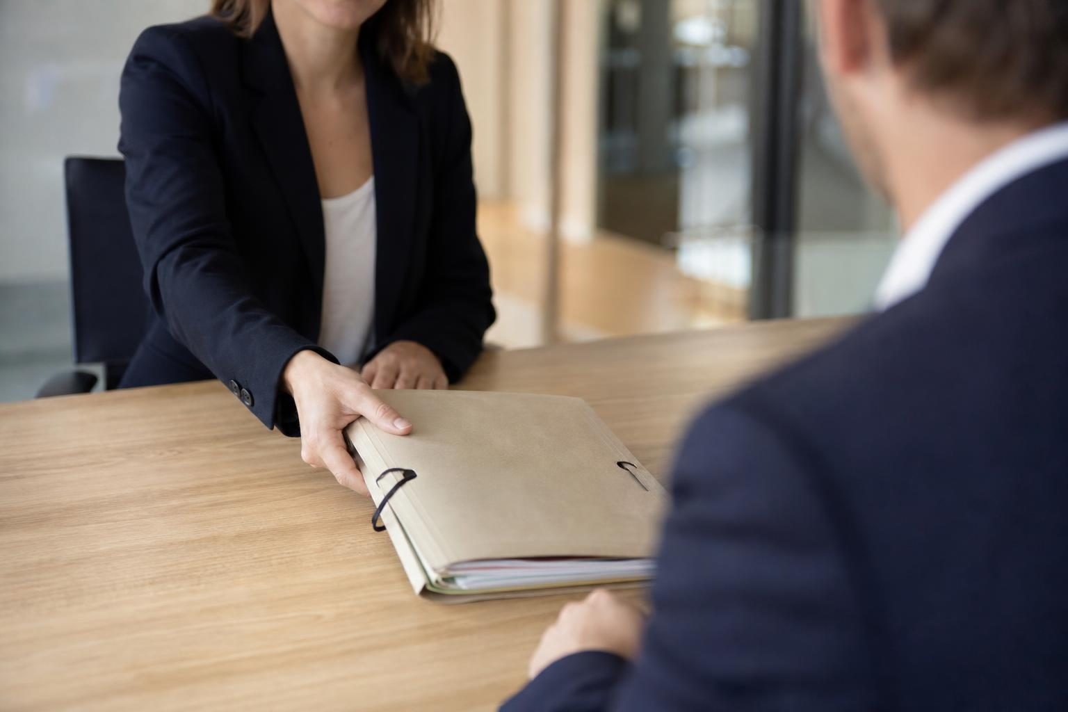 Dans un bureau, une avocate en veste sombre tend un dossier papier à un homme assis en face d’elle.