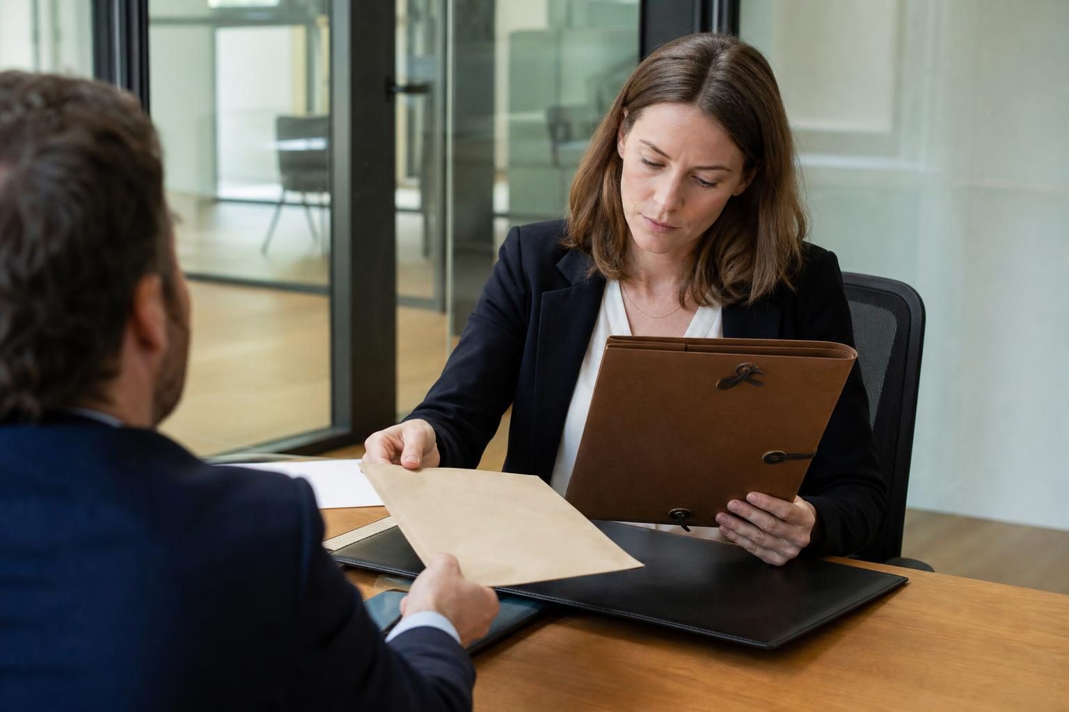 Une avocate en tenue professionnelle examine un dossier en face d’un homme assis à un bureau.