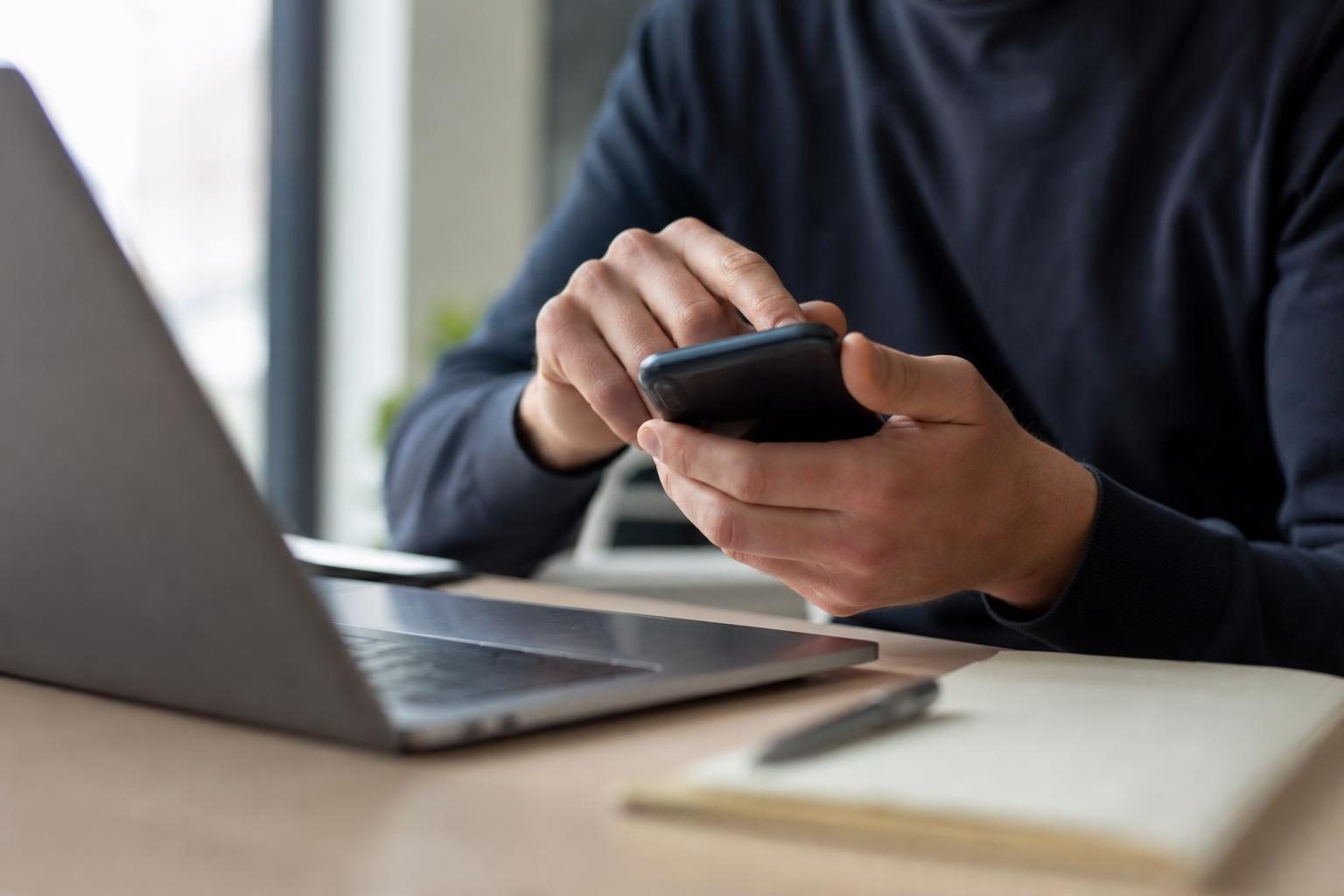 Une personne en pull sombre utilise un smartphone près d’un ordinateur portable posé sur un bureau, avec un carnet et…